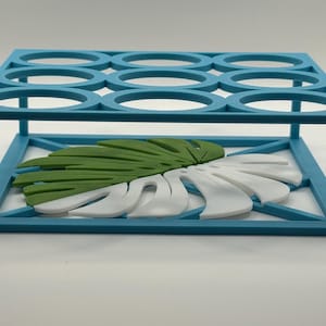 May include: A teal-colored, rectangular rack with nine circular openings on top. A decorative monstera leaf, half green and half white, rests inside the rack. The rack is made of a solid material and is designed to hold items.