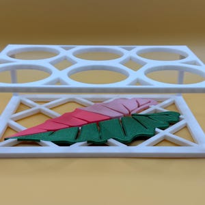 May include: A white, geometric, six-hole holder with a decorative leaf design in pink, red, and green. The holder has a rectangular frame with an open lattice design on the sides.