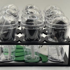 May include: A black, tiered display stand holding clear plastic cups with black contents and clear dome lids. The bottom tier contains clear cups with white and green objects. The stand is on a white surface.