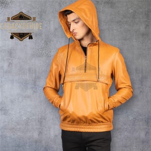 Op de afbeelding: Mosterdgele leren hoodie met een ritssluiting aan de voorkant en twee voorzakken. De jas heeft een capuchon met trekkoord en een geribde zoom. De merknaam "LEGACY OF HIDE" is zichtbaar.