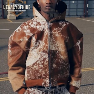 May include: A brown and white cowhide jacket with a zipper closure. The jacket has a hood and brown ribbed cuffs and hem. The image also displays the text "LEGACYOFHIDE" and "Genuine Leather, Jackets, Shirts & Shorts".