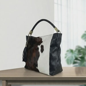 Puede incluir: Un bolso hobo con estampado de piel de vaca en parches marrones, blancos y negros. El bolso tiene una sola asa de cuero negro, un panel lateral de cuero negro y una borla decorativa de cuero negro unida a una cadena de metal dorado.