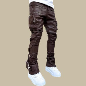 Puede incluir: Pantalones de cuero marrón con múltiples bolsillos y detalles plateados. Los pantalones tienen un diseño apilado en los tobillos y el texto "LEGACYOFHIDE" es visible en la parte trasera. Se usan con zapatillas blancas.