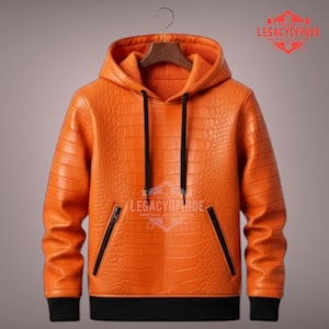Könnte beinhalten: Orangener Leder-Hoodie mit Krokodilhautstruktur. Der Hoodie hat einen Kordelzug, schwarze Akzente und zwei Vordertaschen. Der Saum und die Manschetten sind schwarz. Der Markenname "LEGACYOFHIDE" ist sichtbar.