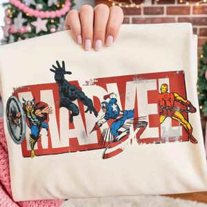 Puede incluir: Camiseta Marvel de color crema con el logotipo de Marvel en rojo desgastado. La camiseta muestra ilustraciones de Thor, Black Panther, Capit&aacute;n Am&eacute;rica e Iron Man en poses de acci&oacute;n. La camiseta est&aacute; sujeta por una persona.