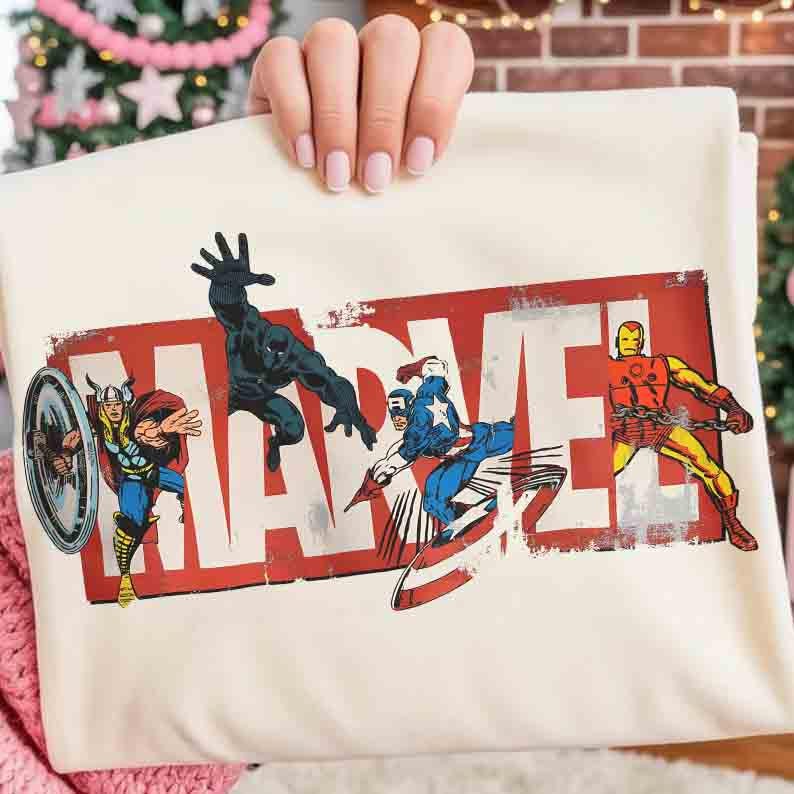 Puede incluir: Camiseta Marvel de color crema con el logotipo de Marvel en rojo desgastado. La camiseta muestra ilustraciones de Thor, Black Panther, Capit&aacute;n Am&eacute;rica e Iron Man en poses de acci&oacute;n. La camiseta est&aacute; sujeta por una persona.