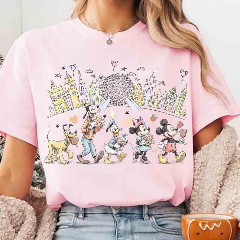 May include: Pink t-shirt featuring a colorful illustration of Disney characters walking in a line, holding food items, with a backdrop of various landmarks. The graphic includes Pluto, Goofy, Donald Duck, Minnie Mouse, and Mickey Mouse.