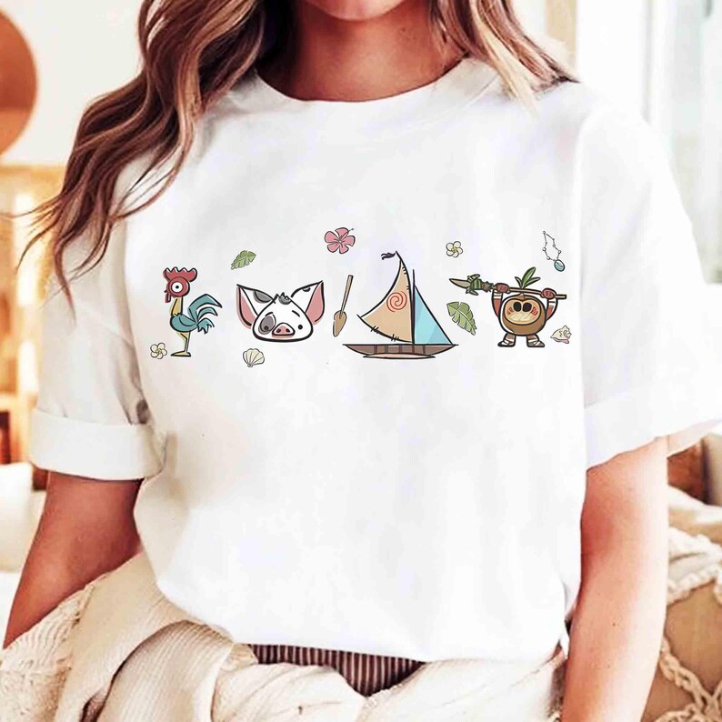 Puede incluir: Una camiseta blanca de manga corta con un estampado gr&aacute;fico horizontal de un gallo de dibujos animados, una cara de cerdo, un remo de madera, un velero con vela en espiral y un peque&ntilde;o personaje de coco sosteniendo una lanza con flores.