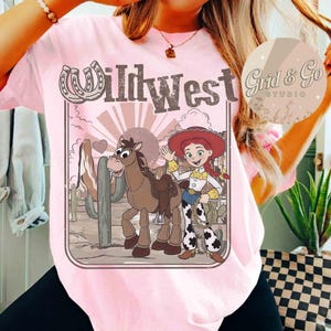 May include: Pale pink t-shirt with a Wild West graphic. The design features a cartoon cowgirl, a horse, and the words "Wild West". The graphic includes a horseshoe, cactus, and a sunburst. The shirt is a light pink colour.