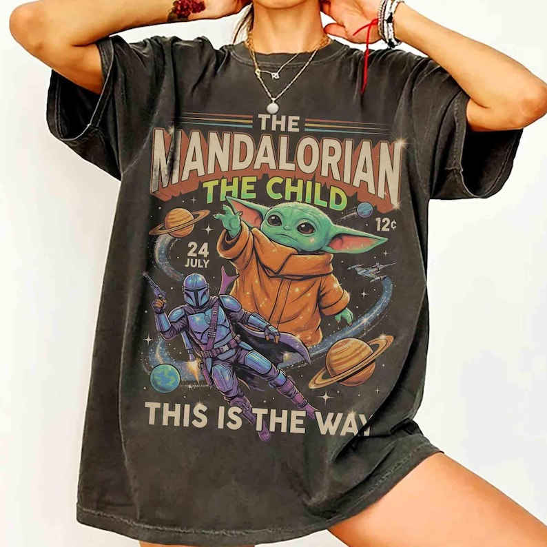 May include: Dark grey t-shirt with a graphic of The Mandalorian and Grogu (The Child). Text reads "The Mandalorian The Child" and "This is the Way". The design includes planets and the date "24 July".
