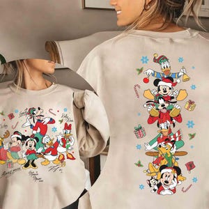 May include: A beige crewneck sweatshirt featuring a graphic print of Disney characters in festive attire. The front displays a group of characters with signatures, while the back shows a vertical stack of characters surrounded by snowflakes and gifts.