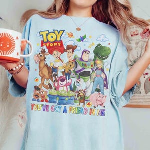 May include: Pale blue t-shirt with a colourful graphic of characters from the film "Toy Story." The design includes Woody, Buzz Lightyear, and other characters, with the text "TOY STORY" and "YOU'VE GOT A FRIEND IN ME".