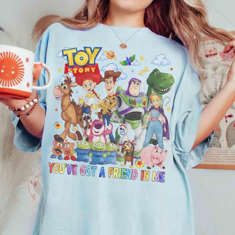 May include: Pale blue t-shirt with a colourful graphic of characters from the film "Toy Story." The design includes Woody, Buzz Lightyear, and other characters, with the text "TOY STORY" and "YOU'VE GOT A FRIEND IN ME".