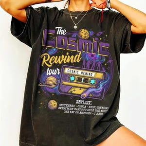 May include: Dark gray t-shirt with a graphic design featuring the text "The Cosmic Rewind Tour" and a purple cassette tape. The design includes planets, stars, and the setlist: September, Conga, Disco Inferno, Everybody Wants to Rule the World, One Way or Another, and I Rain.