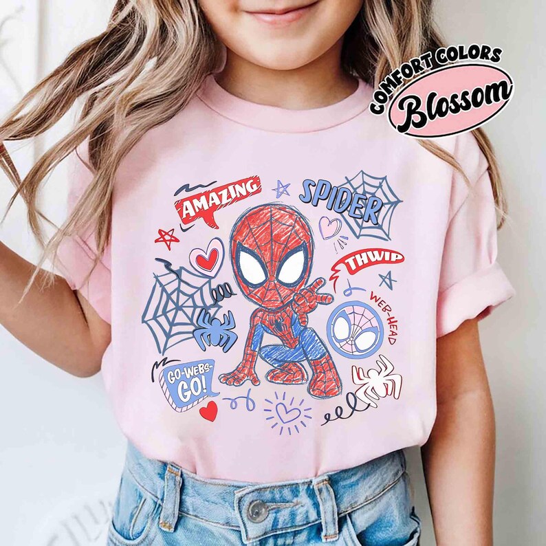 May include: A light pink t-shirt featuring a cartoon Spider-Man design. The graphic includes the words "AMAZING" and "SPIDER", along with spiderwebs, hearts, and the text "GO-WEBS GO!". The shirt is from Comfort Colors, with the word "Blossom" in a pink oval.