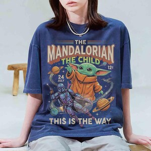 May include: Navy blue t-shirt with a graphic design featuring The Mandalorian and The Child. The design includes the text "The Mandalorian The Child" and "This is the Way", with illustrations of planets and a character in armor.