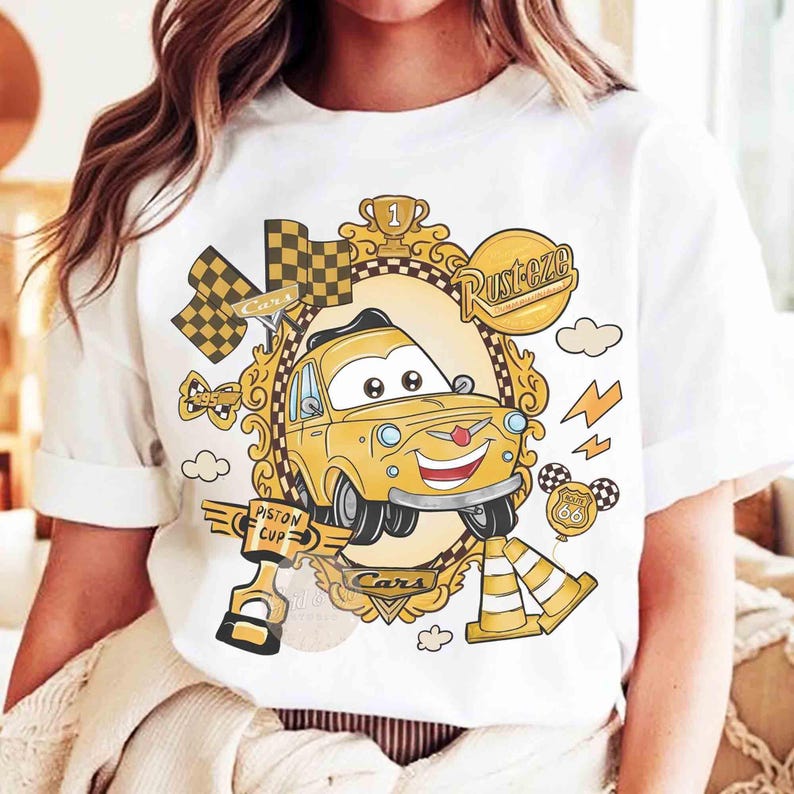 Puede incluir: Camiseta blanca con un dise&ntilde;o de coche de dibujos animados. El coche amarillo tiene una cara sonriente y est&aacute; rodeado de gr&aacute;ficos tem&aacute;ticos de carreras, incluyendo una bandera a cuadros, rayos y las palabras "Cars" y "Piston Cup."
