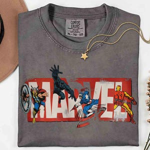 Puede incluir: Camiseta gris Comfort Colors con el logo de Marvel y los personajes de c&oacute;mic Thor, Black Panther, Capit&aacute;n Am&eacute;rica e Iron Man. Un collar con colgante de estrella dorada y un sombrero marr&oacute;n tambi&eacute;n est&aacute;n en la imagen.