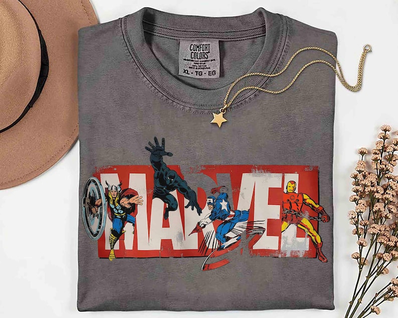 Puede incluir: Camiseta gris Comfort Colors con el logo de Marvel y los personajes de c&oacute;mic Thor, Black Panther, Capit&aacute;n Am&eacute;rica e Iron Man. Un collar con colgante de estrella dorada y un sombrero marr&oacute;n tambi&eacute;n est&aacute;n en la imagen.