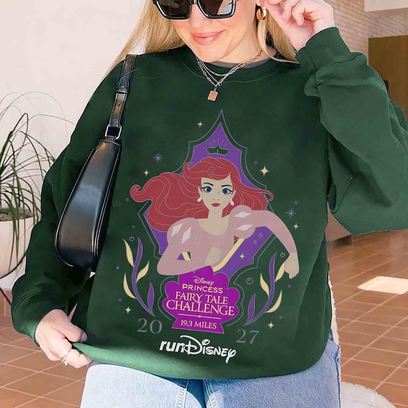 K&ouml;nnte beinhalten: Dunkelgr&uuml;nes Sweatshirt mit einer Grafik von Arielle aus Disneys Der kleinen Meerjungfrau. Die Grafik enth&auml;lt den Text "Disney Princess Fairy Tale Challenge" und "runDisney". Das Sweatshirt hat lange &Auml;rmel und einen Rundhalsausschnitt.