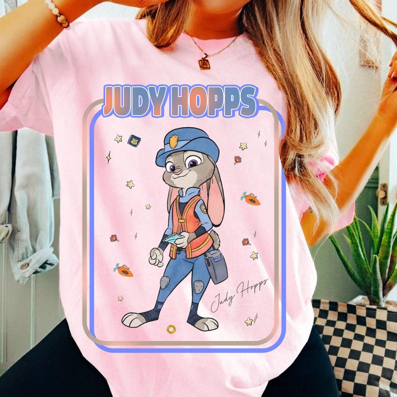 Puede incluir: Camiseta rosa claro con una ilustraci&oacute;n de dibujos animados de Judy Hopps de Zootopia. El dise&ntilde;o presenta a Judy con su uniforme de polic&iacute;a, rodeada de estrellas, zanahorias y el texto "Judy Hopps". La camiseta tiene cuello redondo y mangas cortas.