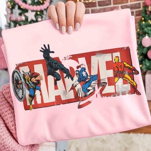 Puede incluir: Una sudadera rosa claro con el logotipo de Marvel en rojo y blanco, con ilustraciones de Thor, Pantera Negra, Capit&aacute;n Am&eacute;rica e Iron Man. La sudadera es sostenida por una persona.