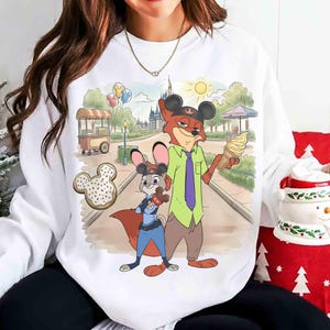 May include: White sweatshirt featuring a cartoon illustration of Nick Wilde and Judy Hopps from Zootopia. The characters are depicted in a park setting with a castle, ice cream, and a Mickey Mouse-shaped donut. The sweatshirt has a crew neck.