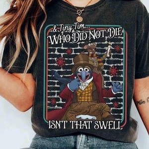 Puede incluir: Camiseta gris oscuro con un gráfico de Gonzo y Rizzo la Rata de Los Muppets, con el texto "& Tiny Tim... WHO DID NOT DIE ISN'T THAT SWELL". El diseño incluye un fondo de pared de ladrillos y copos de nieve.