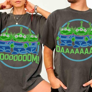 May include: Dark gray t-shirts featuring a cartoon graphic of three green aliens with three eyes each, wearing blue and purple outfits. The text "MOOOOOM!" and "DAAAAAAAD!" are in green. The design is inside a blue circle.