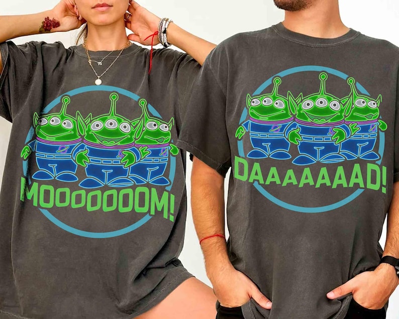 May include: Dark gray t-shirts featuring a cartoon graphic of three green aliens with three eyes each, wearing blue and purple outfits. The text "MOOOOOM!" and "DAAAAAAAD!" are in green. The design is inside a blue circle.