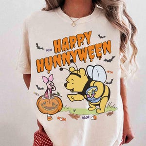 May include: A cream-colored t-shirt featuring a graphic of Winnie the Pooh dressed as a bee and Piglet sitting on a jack-o'-lantern. The text "HAPPY HUNNYWEEN" is printed in orange above the characters, surrounded by small bats and candy icons.