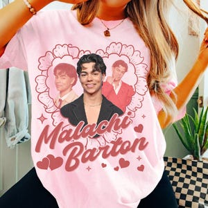 May include: A pale pink t-shirt with a heart-shaped design. The design features three images of a person and the text "Malachi Barton" in a cursive font, along with hearts and stars.