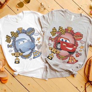 Puede incluir: Dos camisetas con gr&aacute;ficos de coches. Una es blanca con un dise&ntilde;o de coche azul, la otra es gris con un dise&ntilde;o de coche rojo. Cada camiseta presenta un coche de dibujos animados en un marco decorativo.