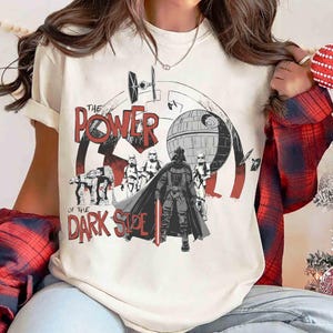 May include: Cream-colored t-shirt featuring a graphic design with the text "THE POWER OF THE DARK SIDE." The design includes Darth Vader, Stormtroopers, and the Death Star, with red and black accents. A Star Wars-themed tee.