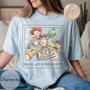 May include: Light blue t-shirt featuring a graphic of Toy Story characters taking a selfie. The image includes Woody, Buzz Lightyear, Jessie, Rex, and other characters. The text "YOU'VE GOT A FRIEND IN ME" is printed below the image.