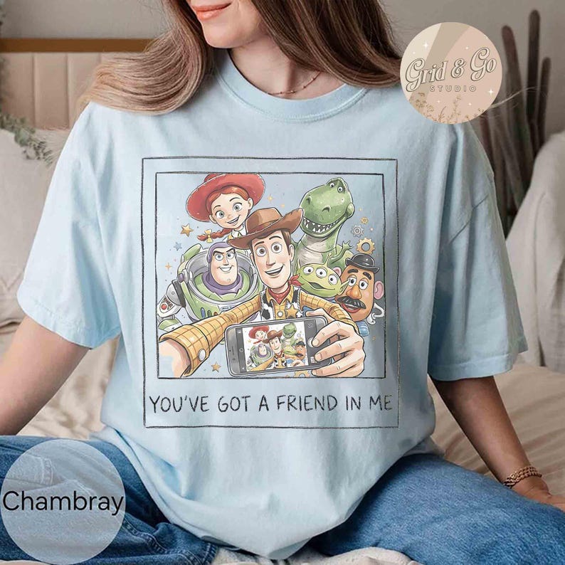 May include: Light blue t-shirt featuring a graphic of Toy Story characters taking a selfie. The image includes Woody, Buzz Lightyear, Jessie, Rex, and other characters. The text "YOU'VE GOT A FRIEND IN ME" is printed below the image.