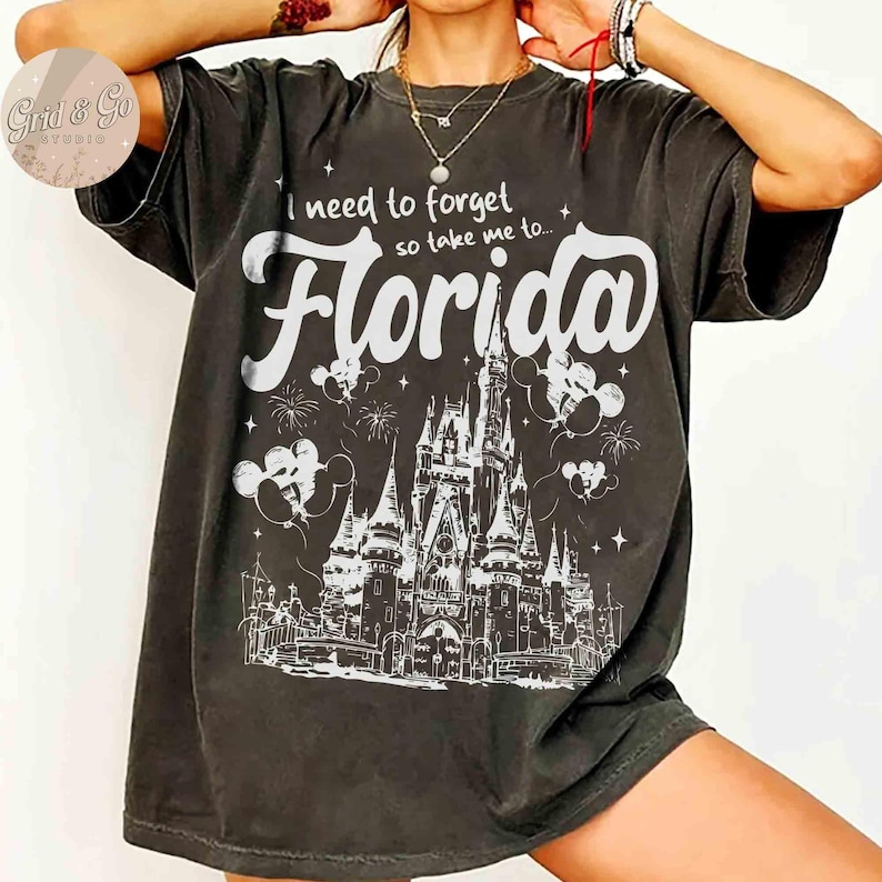 Puede incluir: Camiseta gris oscuro con el texto blanco "I need to forget so take me to... Florida". El dise&ntilde;o incluye una ilustraci&oacute;n detallada de un castillo, fuegos artificiales y siluetas de Mickey Mouse.