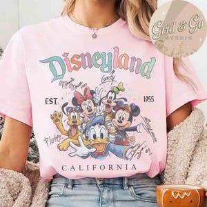 Puede incluir: Camiseta rosa claro con el logo de Disneyland en colores pastel, con Minnie Mouse, Mickey Mouse, Goofy, Donald Duck y Pluto. Incluye el texto "EST. 1955" y "CALIFORNIA".
