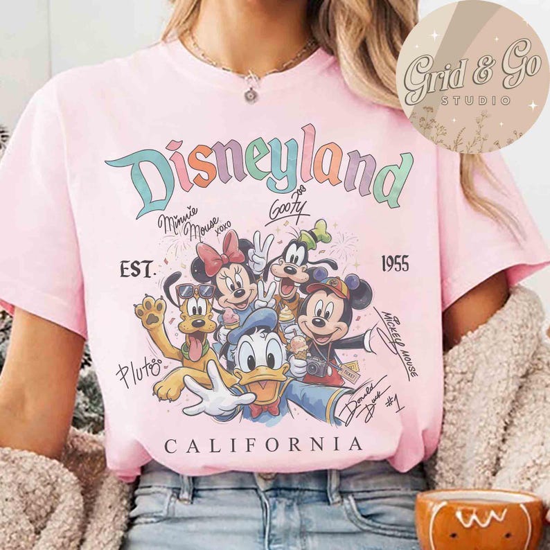 Puede incluir: Camiseta rosa claro con el logo de Disneyland en colores pastel, con Minnie Mouse, Mickey Mouse, Goofy, Donald Duck y Pluto. Incluye el texto "EST. 1955" y "CALIFORNIA".