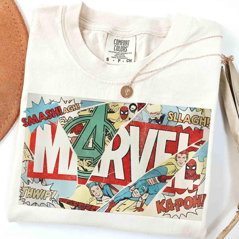 Puede incluir: Camiseta de color crema con un dise&ntilde;o de c&oacute;mic de Marvel. El gr&aacute;fico incluye el logotipo de Marvel, paneles de c&oacute;mic y palabras de acci&oacute;n como "SMASH!" y "KAPOW!". Un collar dorado con un colgante descansa sobre la camiseta.