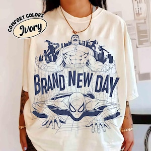 Op de afbeelding: Een ivoorkleurig t-shirt met een donkerblauwe grafische print van de Hulk en Spider-Man met een stadsgezicht. De tekst "BRAND NEW DAY" staat in grote blauwe letters in het midden gedrukt. Een cirkelvormig label vermeldt "COMFORT COLORS IVORY".
