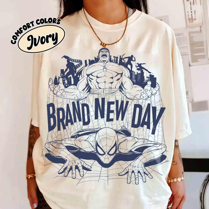 Op de afbeelding: Een ivoorkleurig t-shirt met een donkerblauwe grafische print van de Hulk en Spider-Man met een stadsgezicht. De tekst "BRAND NEW DAY" staat in grote blauwe letters in het midden gedrukt. Een cirkelvormig label vermeldt "COMFORT COLORS IVORY".