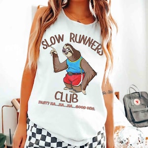 Könnte beinhalten: Weißes Tanktop mit dem Text "SLOW RUNNERS CLUB" und einem Cartoon-Faultier in einem blau-roten Outfit. Der Text "FAST? HA...HA...HA...GOOD ONE." steht unter dem Clubnamen. Das Faultier zeigt nach oben.