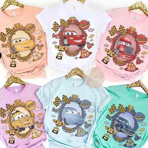 Puede incluir: Colecci&oacute;n de seis camisetas de manga corta en varios colores pastel, cada una con un dise&ntilde;o de coche de dibujos animados dentro de un marco ornamentado. Los dise&ntilde;os incluyen a Rayo McQueen y otros personajes de la franquicia Cars. Las camisetas se muestran en perchas doradas.