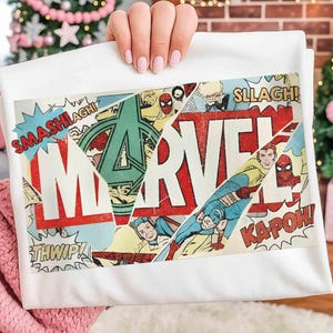 Puede incluir: Camiseta blanca con un dise&ntilde;o de collage de c&oacute;mic de Marvel. El dise&ntilde;o incluye el logotipo de Marvel, vi&ntilde;etas de c&oacute;mic y palabras de acci&oacute;n como "SMASH!" y "KAPOW!" en un estilo vintage.
