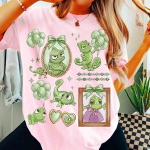 May include: Pink t-shirt featuring a collection of green chameleon illustrations with bows, balloons, and framed portraits. The design includes a heart-shaped locket and decorative stars. The shirt has a crew neck and short sleeves.