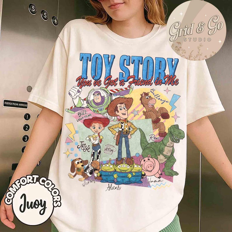 May include: Cream-colored t-shirt featuring a colorful graphic of Toy Story characters, including Woody, Buzz Lightyear, and Jessie. The text "TOY STORY You've Got a Friend in Me" is displayed above the characters. The shirt also has the text "COMFORT COLORS Juoy".