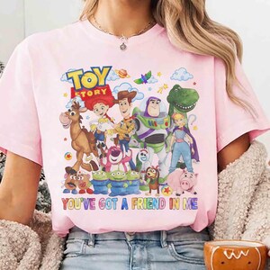 May include: Pale pink t-shirt with a vibrant graphic of "Toy Story" characters. The design includes Woody, Buzz Lightyear, and other characters, with the text "TOY STORY" and "YOU'VE GOT A FRIEND IN ME."