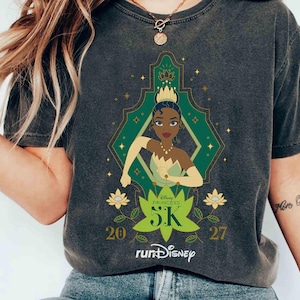 K&ouml;nnte beinhalten: Dunkelgraues T-Shirt mit einer Grafik von Prinzessin Tiana aus Disneys K&uuml;ss den Frosch. Das Design umfasst einen gr&uuml;nen und goldenen geometrischen Rahmen, den Text "Disney Princess 5K" und das runDisney-Logo. Die Zahlen "20" und "27" sind ebenfalls vorhanden.