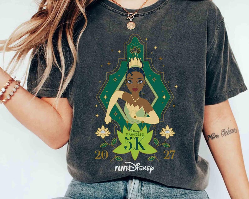 K&ouml;nnte beinhalten: Dunkelgraues T-Shirt mit einer Grafik von Prinzessin Tiana aus Disneys K&uuml;ss den Frosch. Das Design umfasst einen gr&uuml;nen und goldenen geometrischen Rahmen, den Text "Disney Princess 5K" und das runDisney-Logo. Die Zahlen "20" und "27" sind ebenfalls vorhanden.